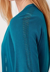 Merenvihreä neulepusero, jossa on V-aukko ja teksturoitu paneeliyksityiskohta hihoissa. Pehmeä kangas ja istuva siluetti.