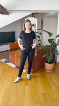 Črna oprijeta majica in črne kavbojke. Srebrni športni čevlji. Notranji prostor z rjavo kordurojno sedežno garnituro in veliko zeleno rastlino v loncu.