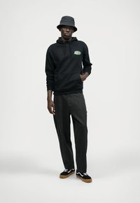 Μαύρη κουκούλα με εμπρόσθια γραφικά, γκρι παντελόνια με κορδόνια και μαύρα αθλητικά παπούτσια με λευκές λεπτομέρειες. Το μοντέλο φοράει γκρι καπέλο του μποξ.