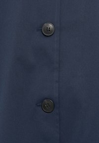 Ύφασμα σε σκούρο μπλε ναυτικού με ομαλή υφή που διαθέτει δύο μαύρα κουμπιά. Ορατές επίπεδες ραφές και απλός, καθαρός σχεδιασμός.