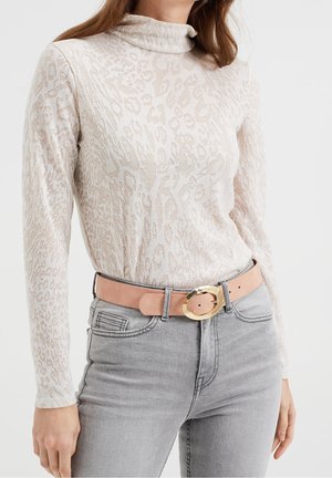 Žena nosí béžový půlprůsvitný top s dlouhým rukávem a zvířecím potiskem, světle šedé džíny a opasek v barvě světle hnědé s velkou zlatou přezkou.