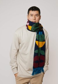 Wełniany szalik z kolorowymi kwiatowymi wzorami na ciemnogranatowym tle, wykończony turkusowymi frędzlami, noszony na jasnobeżowej bluzce z długim rękawem.