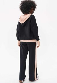 Czarna i różowa bluza z dużym kapturem i długimi rękawami, w zestawie z pasującymi czarnymi spodniami z różowym paskiem po boku. Miękka faktura materiału.