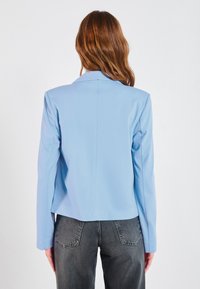 Blazer bleu clair à la coupe ajustée, manches longues et col cranté. Tissu lisse et silhouette épurée se terminant à la taille. Jeans gris visibles.