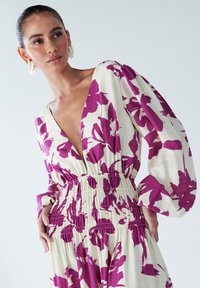 Robe à motifs floraux avec un décolleté en V profond, fond blanc et grandes fleurs violettes. Taille smockée et manches longues évasées.