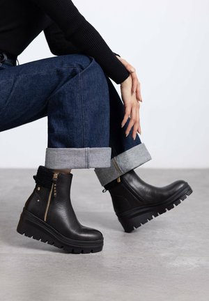 Botas de tobillo de cuero negro con una suela de goma estriada, con un cierre lateral y un acento de lana texturizada en la parte superior, sobre un fondo gris.