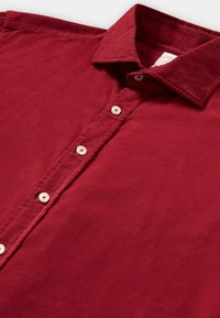 Rode corduroy knoopshirt met witte knopen en een omgeslagen kraag, plat gelegd op een witte achtergrond.