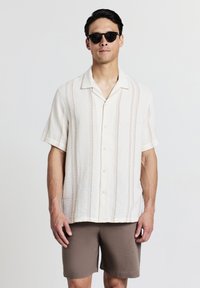 Kurzarmige, hellbeige Bluse mit vertikalen Streifen in sanften Farben und Knopfschluss, kombiniert mit braunen Shorts und schwarzen Sonnenbrillen.