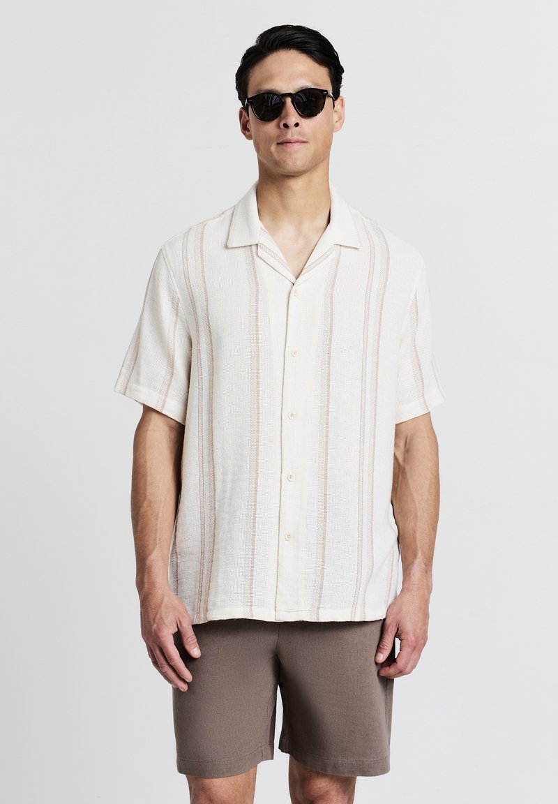 Kurzarmige, hellbeige Bluse mit vertikalen Streifen in sanften Farben und Knopfschluss, kombiniert mit braunen Shorts und schwarzen Sonnenbrillen.