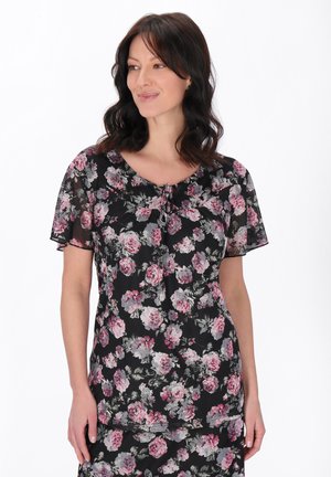 Žena s tmavými vlasy, oblečená v černém květovaném topu s růžovými a šedými květinami, stojící před jednobarevným bílým pozadím.