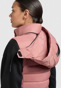Rosa pufferväst med hög krage, med dragkedjefickor och justerbar huva. Texturerat material med en slät yta, lager på ett svart linne.