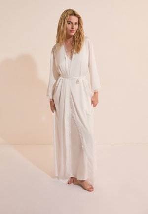 Vrouw met lang blond haar staat op blote voeten en draagt een witte zijden kimono met kanten rand, gestrikt in de taille, tegen een effen beige achtergrond.