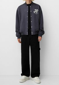 Navy bomber jacket with silver snap buttons, ribbed cuffs, and embroidered logo. Paired with black wide-leg trousers and white sneakers.