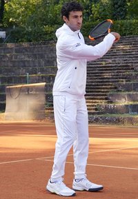 Valge sportlik jakk kapuutsiga, vastavad valged püksid ja valged spordijalatsid. Mängija hoiab tennise reketit, valmistudes teenindama savi väljakul.