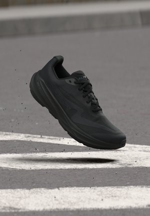 Scarpa da corsa atletica nera sospesa sopra asfalto grigio con segnaletica stradale bianca e piccoli detriti.