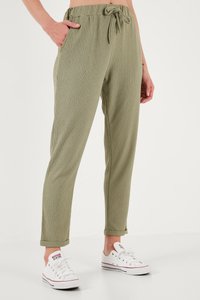 Sieviete valkā olīvu zaļas teksturētas bikses ar uzlocītiem manžetēm un baltas zemas Converse kedas, viena rokā iepīta kabatā, priekšplānā ar vienkrāsainu fonu.