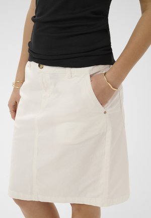 Vrouw draagt een crèmekleurige kokerrok tot op de knie met zakken aan de voorkant en een zwart geribd mouwloos topje, gecombineerd met gouden armbanden en ringen.