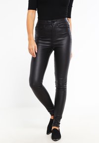 Černé skinny kalhoty z umělé kůže s vysokým pasem, hladkým povrchem a přiléhavým střihem, doplněné černými kotníkovými botami.