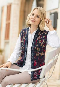 Gebreide tanktop in donkerblauw met bloemen- en paisleypatronen in rood, groen en crème. Gelaagd over een wit overhemd met knoopjes, gecombineerd met lichte broek.
