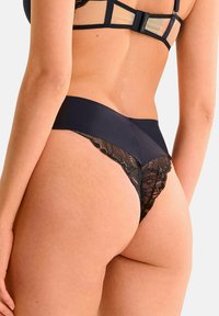 String noir en dentelle et tissu lisse avec une large ceinture élastique, présentant un bord en dentelle festonnée à l'arrière.