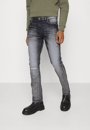 Homme portant un jean noir délavé et usé, des bottes noires épaisses et un haut à manches longues vert olive, debout devant un fond uni.