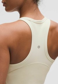 Beżowy sportowy top na ramiączkach z maskującym dekoltem, z logo na plecach oraz gładkiego, elastycznego materiału zapewniającego komfort i swobodę ruchów.