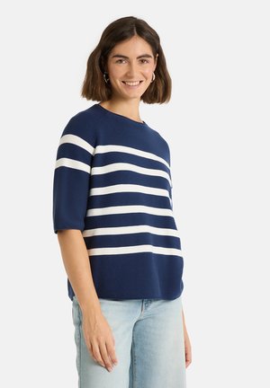 Lächelnde Frau mit schulterlangen braunen Haaren, die einen marineblauen Pullover mit weißen horizontalen Streifen und hellblaue Jeans vor weißem Hintergrund trägt.