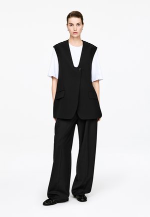 Femme debout portant une chemise blanche à manches courtes, un gilet noir sans manches avec des boutons, un pantalon large noir et des chaussures noires sur fond blanc.