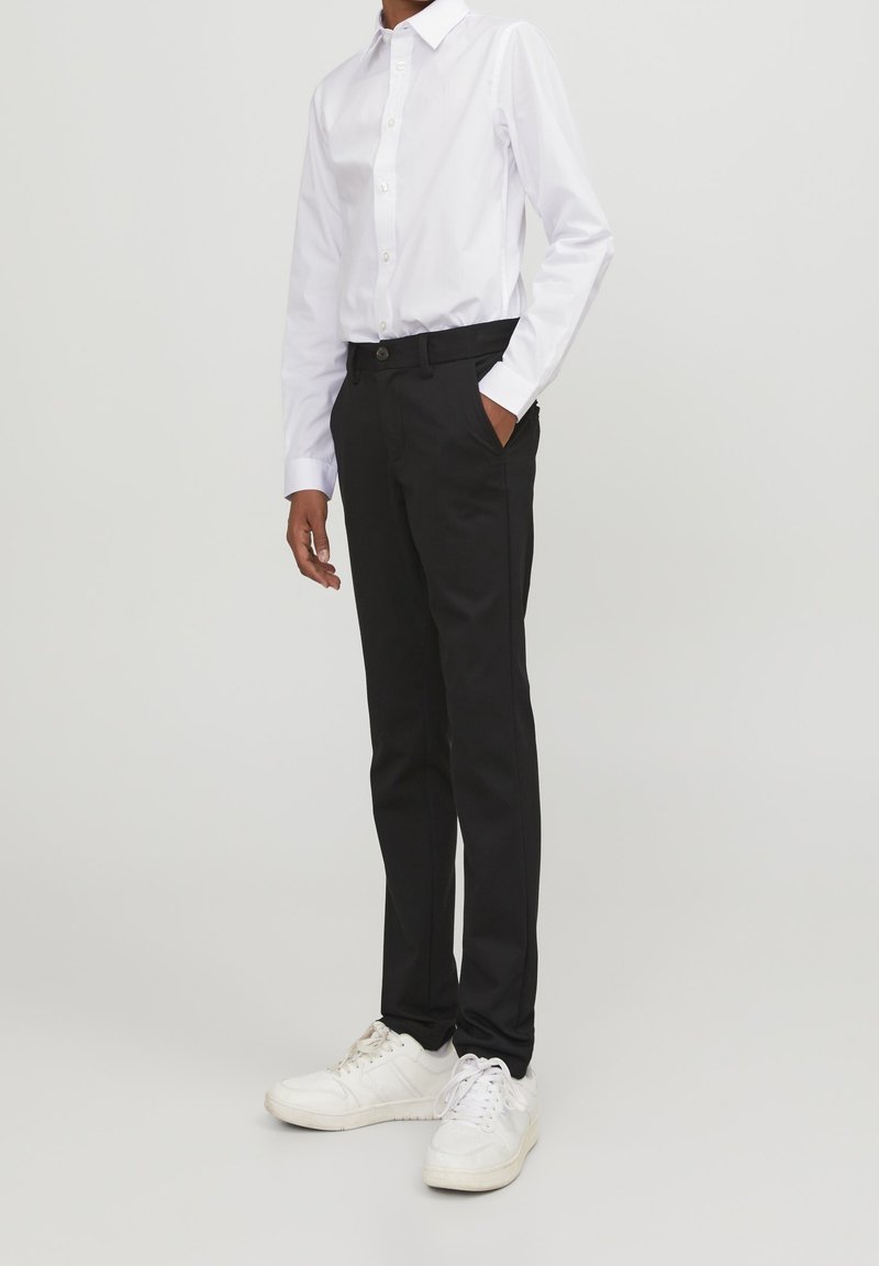 Personne portant une chemise blanche boutonnée, un pantalon noir avec les mains dans les poches, et des baskets blanches, se tenant devant un fond uni.