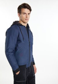 Giacca blu navy con zip frontale, cappuccio nero e polsini a coste. Presenta tasche diagonali frontali e una texture liscia.