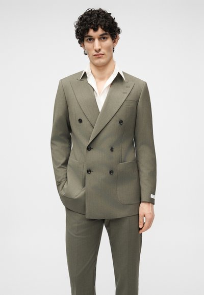 Άνδρας με σγουρά μαλλιά, φοράει διπλόχτυπο χιλαδένιο πράσινο κοστούμι και λευκό πουκάμισο με ανοιχτό γιακά, χέρια στις τσέπες, ουδέτερο φόντο.