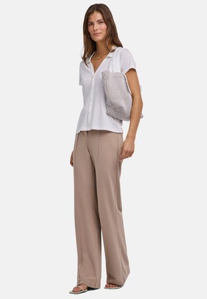 Frau trägt weißes kurzärmeliges Hemd mit Knöpfen, beige Schlaghose, offene Sandalen und hat eine weiße und braune gemusterte Tragetasche über der Schulter hängen.