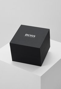 Zwarte kartonnen geschenkdoos met een scharniereksel, met in reliëf "BOSS" en "HUGO BOSS" in zilveren letters, vierkante vorm, gladde textuur.