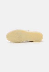 Sneaker outsole in yellow rubber with a textured pattern, featuring a circular logo and brand name. Smooth and rounded edges.