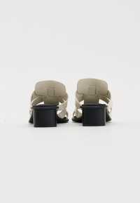 Sandales à talons beiges avec des straps croisées ; talon carré noir ; matière lisse ; design à dos ouvert ; détails minimalistes.