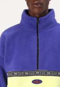 Lila fleece pulóver magas gallérral és fekete cipzárral. Mintás fekete sávval és világos sárga alsó résszel rendelkezik, amelyen logó folt található.