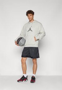 Grå hættetrøje med kengurulomme, sorte shorts med mønster og røde detaljer på sorte sportssko. Holder en struktureret basketball.