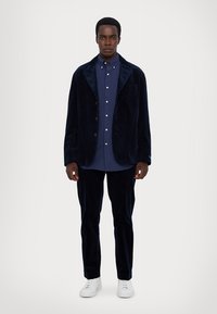 Marineblå fløyelsblazer med klaffelapper over en blå prikkete skjorte. Kombinert med matchende bukser og hvite sneakers. Teksturerte stoffdetaljer.