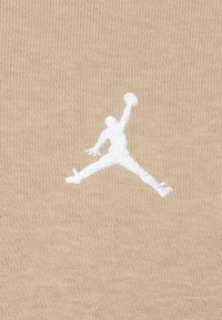 Tissu beige clair avec une texture douce mettant en avant un logo Jumpman brodé en blanc au centre du matériau. Design minimaliste sans accents supplémentaires.