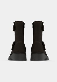 Botines de cuero negro con suela gruesa, con hebillas y cremalleras laterales para facilitar su uso. Acabado texturizado y diseño minimalista.