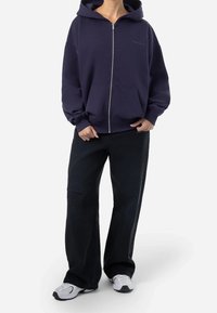 Felpa a zip navy con tasche anteriori, abbinata a jeans neri a gamba larga con dettagli strappati, e scarpe sportive bianche.