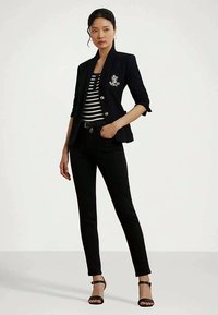 Γυναίκα που στέκεται φορώντας μαύρο σακάκι με λευκή κεντημένη διακόσμηση, μαύρο και άσπρο ριγωτό τοπ, μαύρο παντελόνι και μαύρες ψηλές σαγιονάρες.