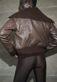 Giubbotto bomber in pelle marrone con un ampio colletto a coste e polsini. La texture appare liscia con un orlo aderente. Sotto vengono indossati leggings neri.