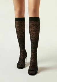Schwarze kniehohe Socken mit floralem Spitzenmuster, ausgestattet mit einem festen schwarzen Bündchen an der Oberseite. Getragen mit schwarzen offenen High Heels.