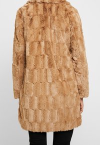 Beige pälsjacka i fuskpäls med ett strukturerat fyrkantigt mönster, utrustad med en bred krage och en lös passform. Jackan sträcker sig till mitten av låret.