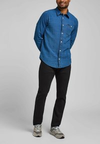 Camisa azul de botones con un patrón texturizado, pantalones negros y zapatillas grises. La camisa tiene un bolsillo en el pecho y un dobladillo curvado.