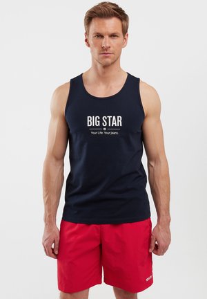 Tumesinine tank top valge "BIG STAR" graafika ja tekstiga. Punased lühikesed püksid, kerge kangas. Igapäevane suvine riietus.