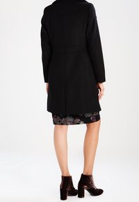 Svart ullkappa med rak skärning, synliga sömdetaljer och slagkrage, kombinerad med en blommönstrad kjol och bruna ankelskor.