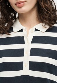 Γυναίκα με μπούκλες, φορώντας μπλε ναυτικό-λευκό ριγέ πουκάμισο με γιακά και κουμπιά μπροστά, καθώς και μικρό κεντημένο λογότυπο.