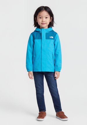 Παιδί που στέκεται προς τα εμπρός φορώντας μπλε μπουφάν με κουκούλα, σκούρο τζιν και καφέ αθλητικά παπούτσια σε λευκό φόντο.